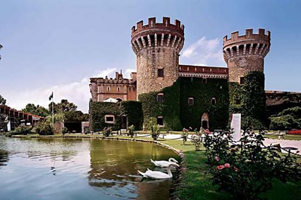 Eine Burg mit zwei Türmen steht umgeben von üppigem Grün und einem See. Schwäne schwimmen im Wasser, während Menschen im Garten spazieren. Es ist ein klarer, sonniger Tag.
