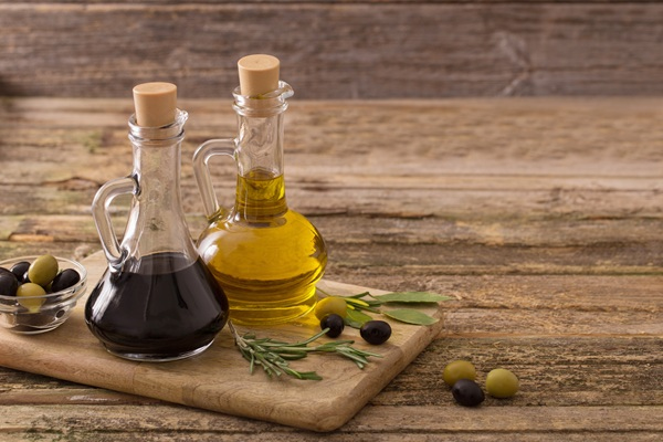 Zwei Glaskaraffen stehen auf einem Holzbrett. Die linke enthält dunkles, dickflüssiges Öl, die rechte helles, flüssiges Öl. Daneben liegen grüne und schwarze Oliven sowie frische Rosmarinzweige.