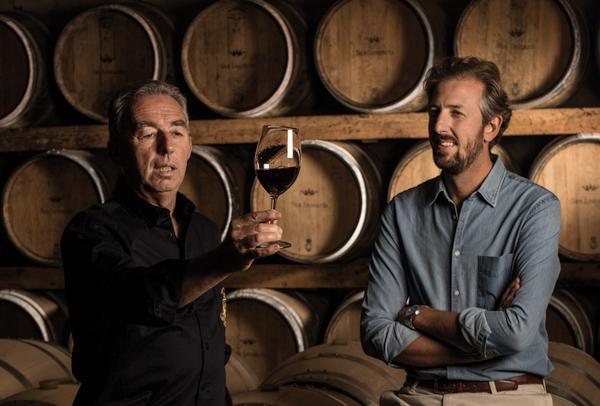Ein Mann hält ein Weinglas mit Rotwein in die Höhe, während ein anderer Mann daneben steht und zusieht. Im Hintergrund sind viele Holzfässer zu sehen, was auf eine Weinlagerung hinweist.