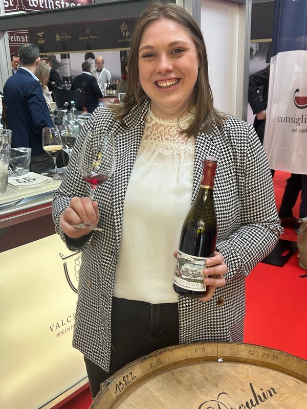 Eine lächelnde Frau hält ein Weinglas in einer Hand und eine vinierte Flasche in der anderen. Im Hintergrund sind Weinbarren und Messestände sichtbar, was eine Weinausstellung andeutet.