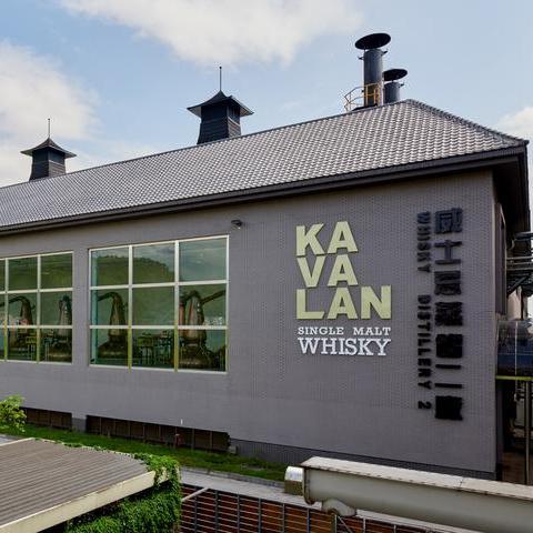 Ein modernes Whisky-Destillerie-Gebäude mit großen Fenstern, durch die Kupfer destillierbehälter sichtbar sind. Das Gebäude hat ein graues Fassadenfinish und das Logo -KAVALAN SINGLE MALT WHISKY- ist prominent angebracht.