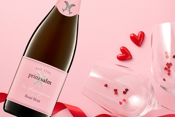 Eine Flasche Roséwein steht auf einem rosa Hintergrund. Zwei Gläser mit Herzchen und rote Herzchen sind verstreut, um eine romantische Stimmung zu erzeugen. Ein rotes Band liegt daneben.