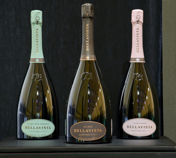 Drei Flaschen Schaumwein stehen nebeneinander auf einem schwarzen Untergrund. Die Flaschen haben unterschiedlich gefärbte Kappen: grün, braun und rosa. Jede Flasche trägt eine Etikette mit dem Namen -BELLAVISTA-.