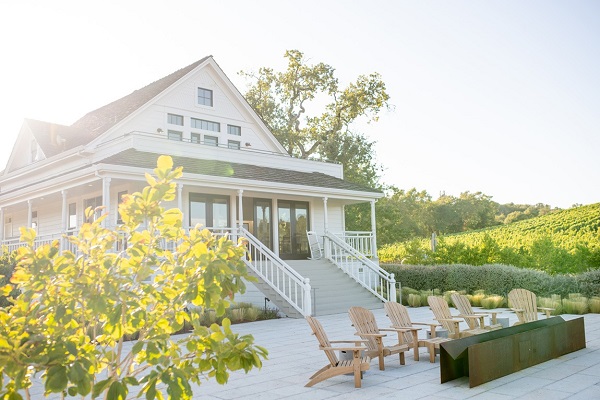 Ein großes, helles Gebäude mit einer Veranda ist im Hintergrund zu sehen. Vor dem Gebäude stehen mehrere Holzstühle auf einem gepflasterten Bereich, umgeben von Weinbergen. Die Sonne scheint.