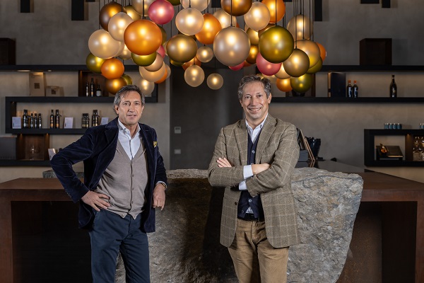 Zwei Männer stehen vor einem großen, grauen Stein an einer modernen Bar. Über ihnen hängen dekorative, bunte Lampen in verschiedenen Größen. Im Hintergrund sind Regale mit Flaschen sichtbar.