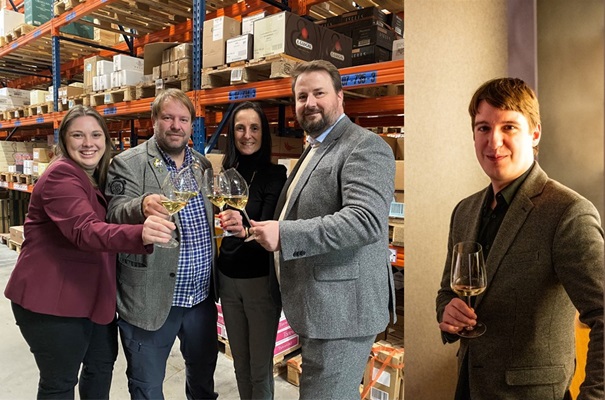Eine Gruppe von vier Personen in Anzügen hält Weingläser und stößt an, während sie in einem Lager mit Regalen voller Kartons stehen. Rechts steht eine weitere Person alleine mit einem Weinglas in der Hand.
