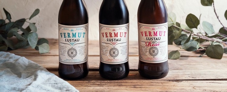 Drei Flaschen Vermut Lustau stehen auf einem Holztisch. Sie sind unterschiedlich gefärbt: eine grüne, eine schwarze und eine rosafarbene Flasche. Im Hintergrund sind grüne Blätter sichtbar.