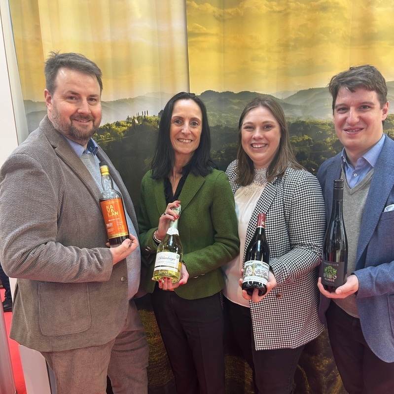 Vier Personen stehen nebeneinander und halten jeweils eine Flasche. Sie wirken freundlich und posierend vor einem Hintergrund mit einer landschaftlichen Darstellung. Die Atmosphäre ist professionell und einladend.