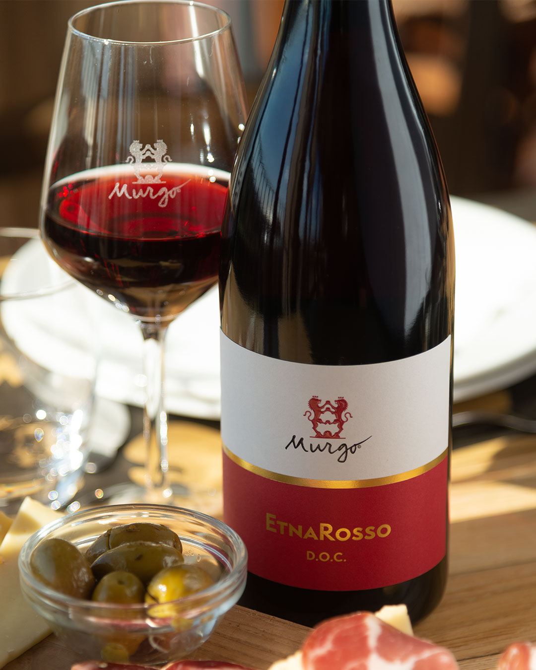 Rotwein in einem Glaspokal und einer Flasche mit etikettierter -EtnaRosso D.O.C.--Beschreibung. Daneben eine Schale mit Oliven und Wurst auf einem Holzbrett. Helle, einladende Umgebung.