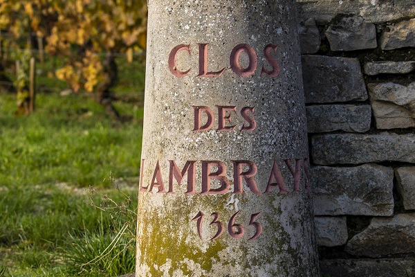 Ein steinerner Pfosten mit der Inschrift -CLOS DES LAMBRAY 1365- steht in einem Weinberg, umgeben von grünen Wiesen und Weinstöcken im Hintergrund.