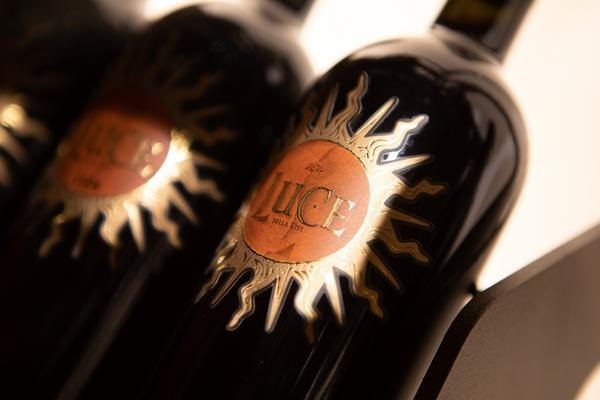 Dunkle Weinflaschen stehen in einer Reihe, mit einem goldenen Etikett, das die Worte -LUCE- und eine Sonnensymbolik zeigt. Der Hintergrund ist verschwommen und neutral.