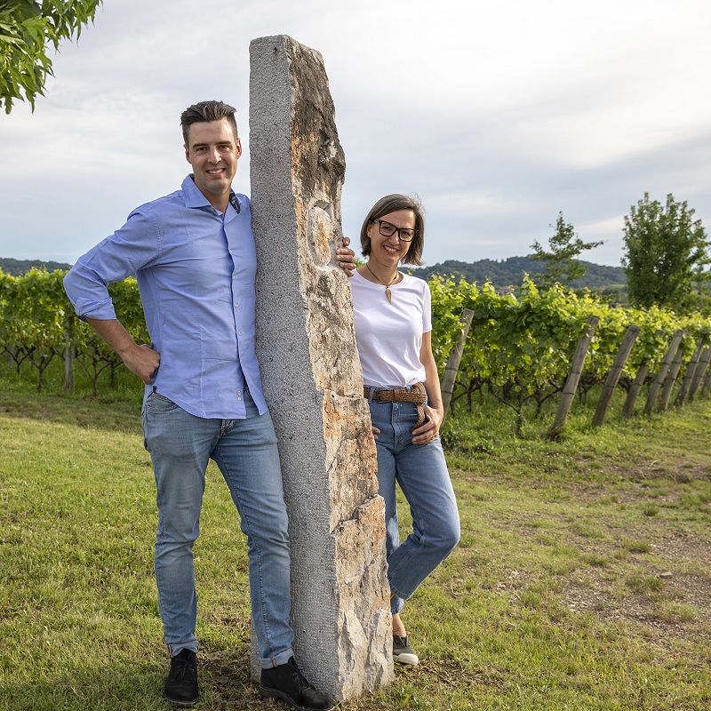 Zwei Personen stehen lächelnd neben einem großen Stein in einem Weinberg. Im Hintergrund sind Weinreben und ein leicht bewölkter Himmel sichtbar.