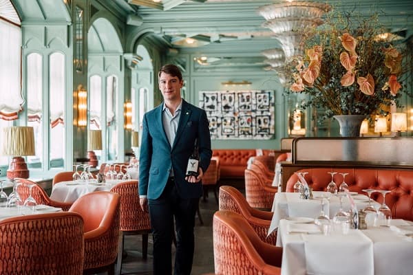 Ein junger Mann in einem eleganten blauen Blazer steht in einem stilvollen Restaurant mit grünen Wänden. Rote gepolsterte Stühle und weiße Tischdecken schaffen eine gehobene Atmosphäre. Obst- und Blumendekoration vervollständigt die Umgebung.