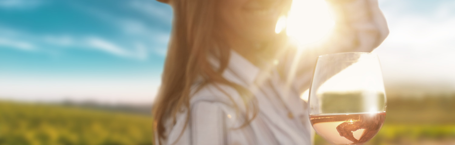 Eine Person hält ein Weinglas mit Roséwein in der Hand, während sie in Richtung der Sonne lächelt. Im Hintergrund erstrecken sich Weinberge unter einem blauen Himmel mit leichten Wolken.