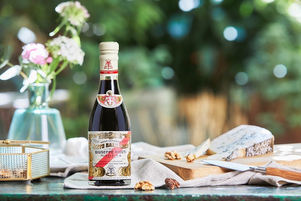 Eine Flasche Balsamico-Essig steht auf einem Tisch, umgeben von Blumen, Nüssen und einem Stück Käse. Im Hintergrund sind unscharfe grüne Pflanzen zu sehen, die eine sommerliche Atmosphäre schaffen.
