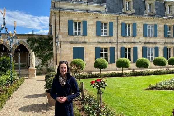 Eine Frau steht im Vordergrund eines gepflegten Gartens, umgeben von grünen, runden Büschen und einer blühenden Rosenpflanze. Im Hintergrund sieht man ein großes, historisches Gebäude mit Fenstern.