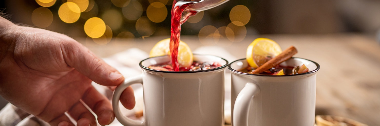 Zwei rote Becher mit warmem Getränk sind gefüllt, dekoriert mit Orangenscheiben und Zimtstangen. Getrocknete Orangenscheiben und Gewürze liegen daneben, umgeben von herbstlichen Blättern.
