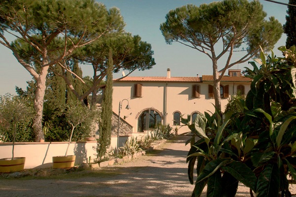 Ein großes, helles Gebäude mit Fensterläden steht im Mittelpunkt, umgeben von hohen Pinien und grünen Pflanzen. Ein geschotterter Weg führt zum Eingang des Hauses, das in einer ländlichen Umgebung liegt.
