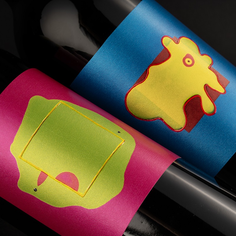 Zwei Flaschen mit bunten Etiketten sind abgebildet. Die Flasche links hat ein pinkes Etikett mit einer grünen, geometrischen Form und einer kleinen roten Fläche. Die rechte Flasche trägt ein blaues Etikett mit einer gelben, organischen Form.