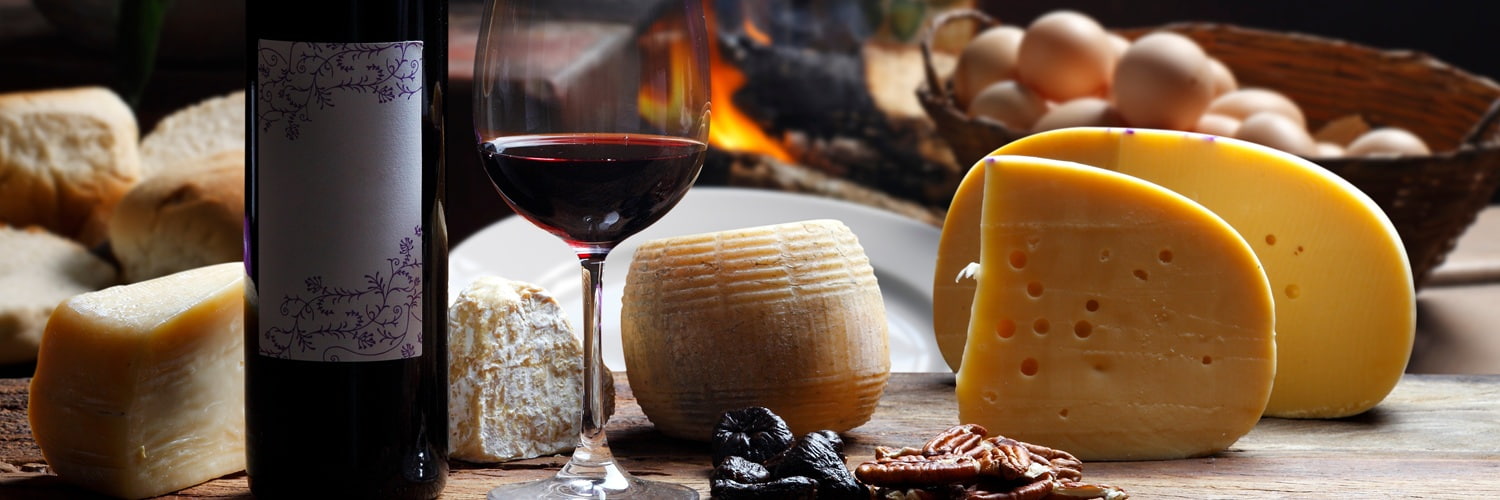 Ein Holzbrett zeigt eine Auswahl an Käsesorten und einen Rotwein in einem Glas. Dazu liegen ein Käsemesser und Trauben sowie ein Apfel. Der Hintergrund ist leicht strukturiert.