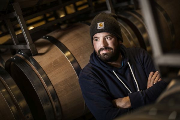 Ein Mann mit Bart trägt einen dunkelblauen Kapuzenpullover und eine Mütze. Er lehnt entspannt an einem Regal mit großen Holzfässern in einem Weinkeller.