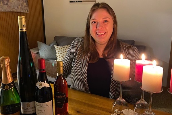 Eine lächelnde Frau sitzt an einem Tisch, umgeben von verschiedenen Weinen und Kerzen. Im Hintergrund ist ein Sofa sichtbar, das eine gemütliche Umgebung schafft.