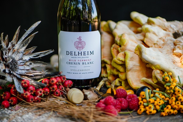 Eine Flasche Delheim Chenin Blanc steht im Vordergrund, umgeben von frischem Obst wie Himbeeren, kleinen roten Beeren und einem Ananasblatt. Im Hintergrund liegen Pilze und dekorative Blumen.