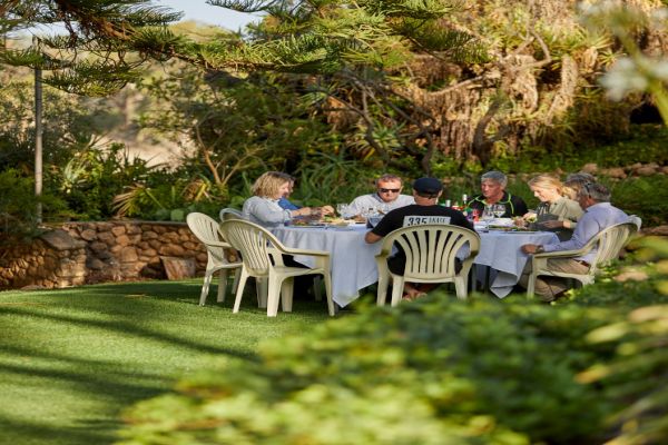 Eine Gruppe von sechs Personen sitzt an einem runden Tisch im Freien und isst. Um sie herum ist ein gepflegter Garten mit grünem Rasen und Bäumen im Hintergrund.