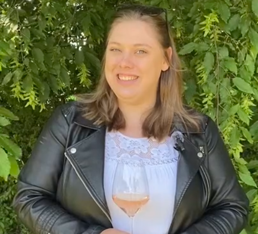 Junge Frau mit brünettem Haar in einer Lederjacke hält ein Glas Roséwein und lächelt, während sie vor einem Hintergrund aus grünen Blättern steht.
