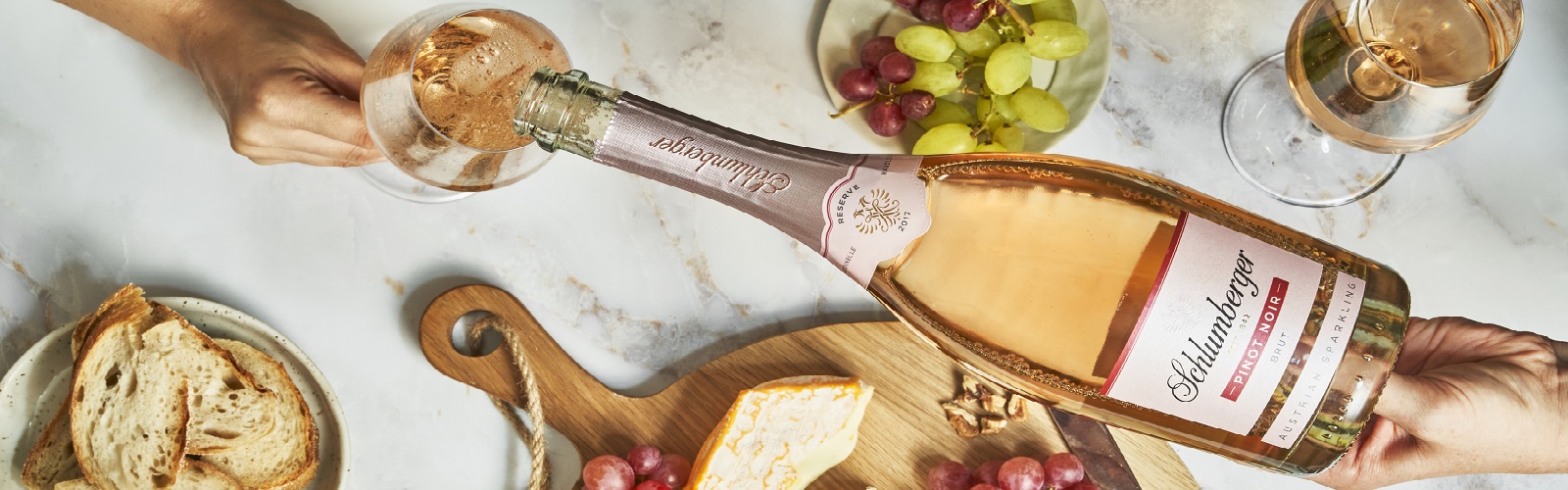 Schlumberger Sektkellerei Eine Flasche Schlüterberg Sekt wird in ein Glas eingeschenkt. Im Hintergrund liegen Brot, Käse, Trauben und eine weitere Glas mit Sekt auf einem Marmortisch.