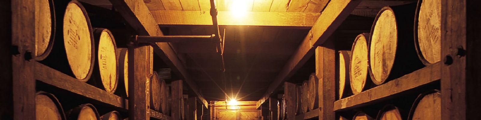 Blanton's Fässer aus Holz sind auf Regalen in einem schwach beleuchteten Lagerraum angeordnet. Ein warmes Licht leuchtet von der Decke und schafft eine gemütliche Atmosphäre.