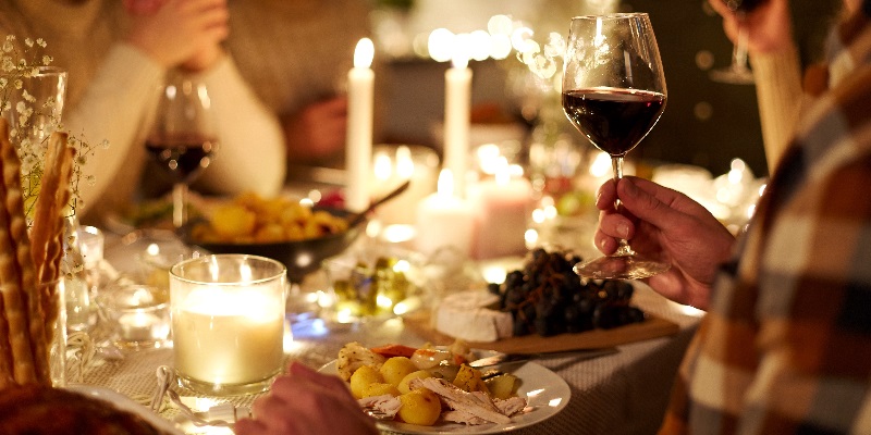 Ein gedeckter Tisch mit Kerzenlicht und festlichem Essen. Eine Person hält ein Weinglas, während andere um den Tisch sitzen. Es gibt verschiedene Speisen, darunter Kartoffeln, Trauben und Käse.
