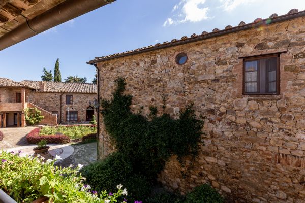 Ein steinernes Gebäude mit einem runden Fenster und grünem, kletterndem Pflanzenbewuchs steht im Vordergrund. Im Hintergrund sind weitere Häuser und eine blühende Gartenlandschaft zu sehen, unter blauem Himmel.