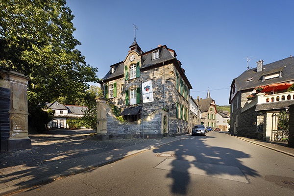 Ein historisches Gebäude aus grauem Stein steht an einer Straßenkreuzung. Es hat mehrere Fenster und ein Schild. Es ist ein sonniger Tag mit Bäumen im Hintergrund. Ein Auto fährt vorbei.
