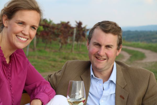 Ein Mann und eine Frau sitzen lächelnd zusammen, umgeben von Weinreben. Der Mann hält ein Glas Weißwein, während die Frau eine lila Bluse trägt. Der Hintergrund zeigt eine hügelige Landschaft.