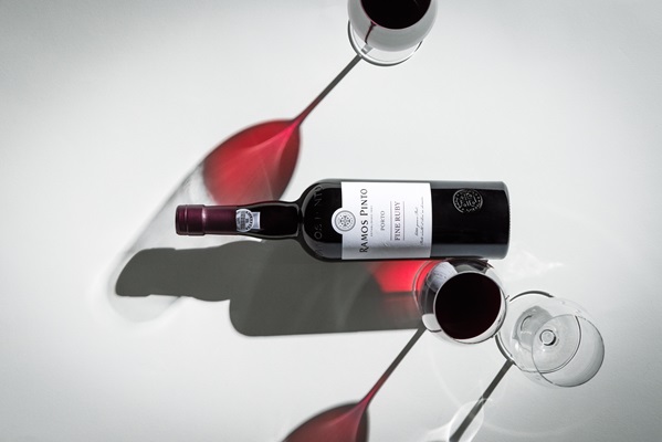 Eine Flasche Rotwein liegt auf einem weißen Tisch, umgeben von Weingläsern, aus denen der Wein schimmert. Die Schatten des Weins und der Gläser erzeugen ästhetische Formen.