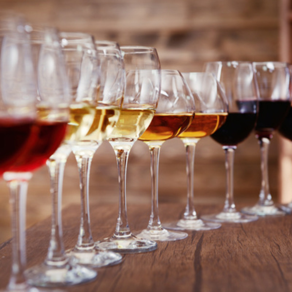 Eine Reihe von Weingläsern steht auf einem Holztisch. Die Gläser enthalten verschiedene Weinsorten in unterschiedlichen Farbtönen, von rot über weiß bis hin zu rosé.