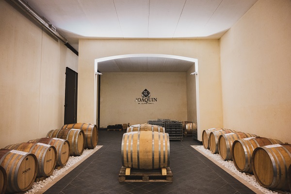 Holzfässer sind in einem lichtdurchfluteten Raum angeordnet, mit einem zentralen Durchgang. Der Boden ist mit dunklen Fliesen und dekorativem Kies versehen. An der Wand ist ein Logo sichtbar.