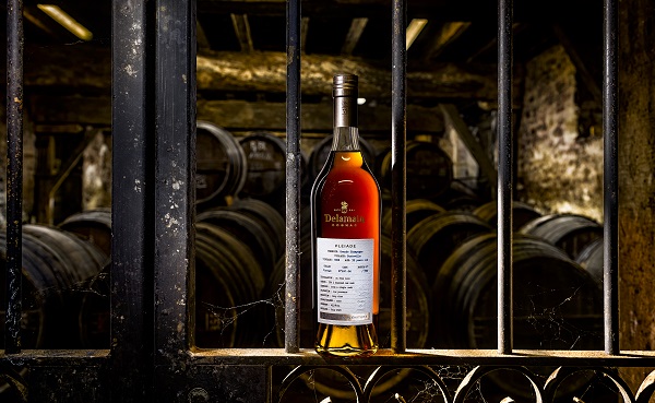 Eine Flasche Delamain Cognac steht auf einem Regal vor einem Hintergrund aus Holzfässern. Das Licht betont die goldene Flüssigkeit in der Flasche. Es herrscht eine rustikale Atmosphäre, die an eine alte Lagerstätte erinnert.