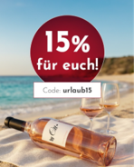 Eine Flasche Roséwein liegt auf einem Handtuch am Strand, daneben zwei Weingläser. Im Hintergrund ist das Meer und die Küste sichtbar. Text: -15% für euch! Code: urlaub15-.