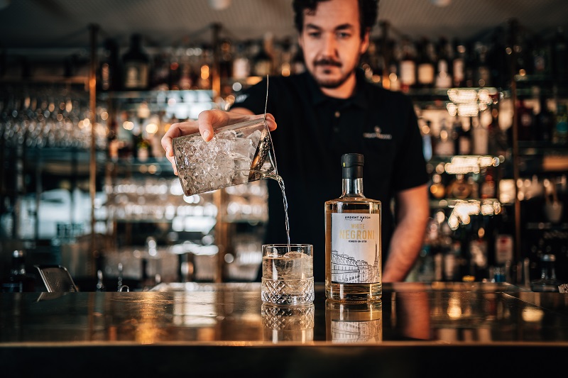 Ein Barkeeper gießt Eis und einen klaren Drink in ein Glas auf einer glänzenden Theke, während eine Flasche mit der Aufschrift -White Negroni- im Vordergrund steht. Im Hintergrund sind Regale mit Spirituosen sichtbar.