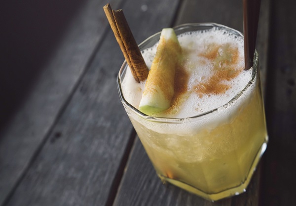 Ein Glas mit einem schaumigen Getränk steht auf einem Holzuntergrund. Zwei Zimtstangen und eine Melonenscheibe verzieren das Getränk. Es wirkt frisch und einladend.