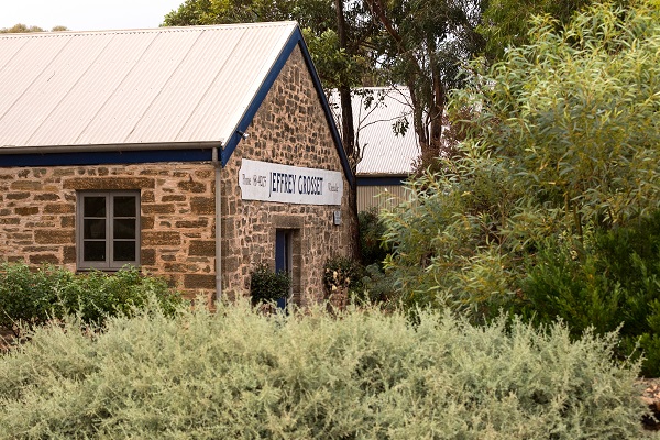 Ein steinernes Gebäude mit einem blauen Dach und Fensterrahmen, das ein Schild mit -Jeffrey Grosse- zeigt. Im Vordergrund wächst grünes Gestrüpp und Sträucher.