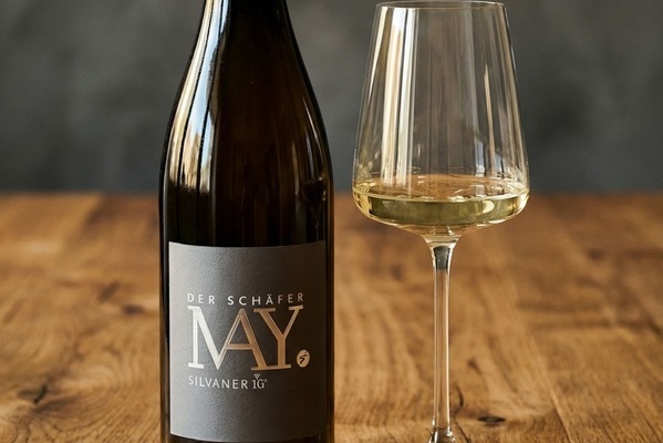 Eine Flasche Silvaner-Wein steht neben einem Weinglas, das mit hellgelbem Wein gefüllt ist. Die Flasche hat ein elegantes, graues Etikett mit dem Schriftzug -DER Schäfer MAY-. Der Hintergrund ist unscharf.