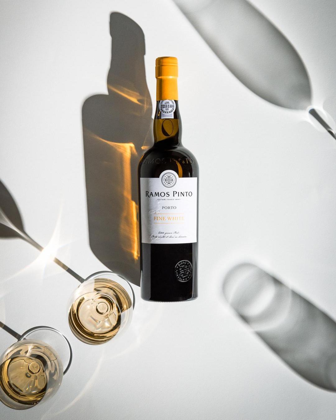 Eine Flasche Ramos Pinto Porto, Fine White, steht auf einer weißen Oberfläche. Darunter liegen zwei Gläser mit hellgelbem Wein, während Schatten und Lichtverhältnisse interessantes Spiel erzeugen.
