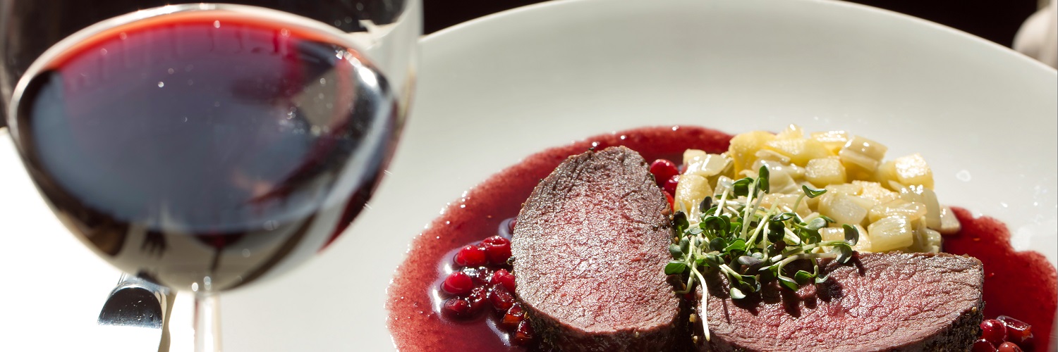 Der Sellerietrick: So wird alter Wein wieder munter Ein Glas Rotwein steht im Vordergrund. Im Hintergrund liegt ein kunstvoll angerichteter Teller mit zwei Scheiben Fleisch in einer Sauce, umgeben von kleinen Beilagen und frischen Kräutern.