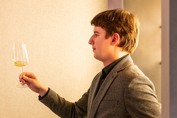 Ein junger Mann steht seitlich, hält ein Weinglas mit hellem Wein in der Hand und blickt nachdenklich auf das Glas. Der Hintergrund ist hell und strukturiert.