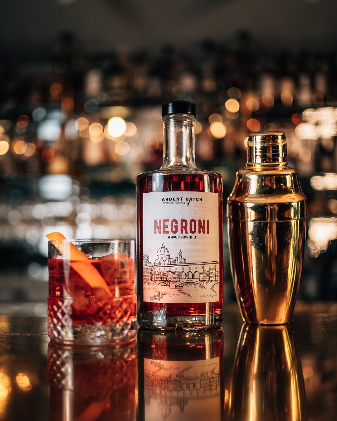 Ein Flasche Negroni mit einem dekorativen Etikett steht neben einem Cocktail-Shaker aus Gold und einem Glas mit einem roten Drink, garniert mit Orangenschale, auf einer glänzenden Baroberfläche.