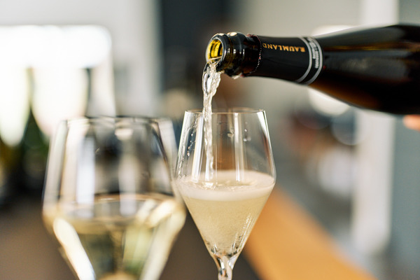 Ein Sekt wird aus einer Flasche in ein dünnwandiges Glas gegossen, während ein zweites Glas daneben steht. Der Hintergrund ist unscharf und zeigt eine helle Umgebung.