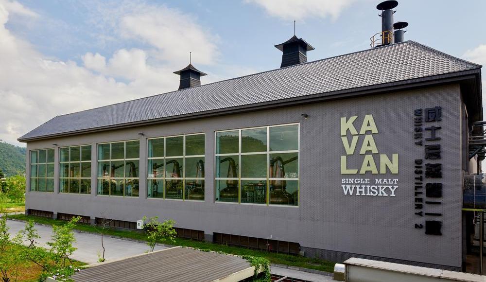 Ein modernes Gebäude mit großen Fenstern zeigt eine Whisky-Destillerie. Darauf steht -KAVALAN SINGLE MALT WHISKY-. Im Hintergrund sind sanfte Hügel und ein blauer Himmel sichtbar.